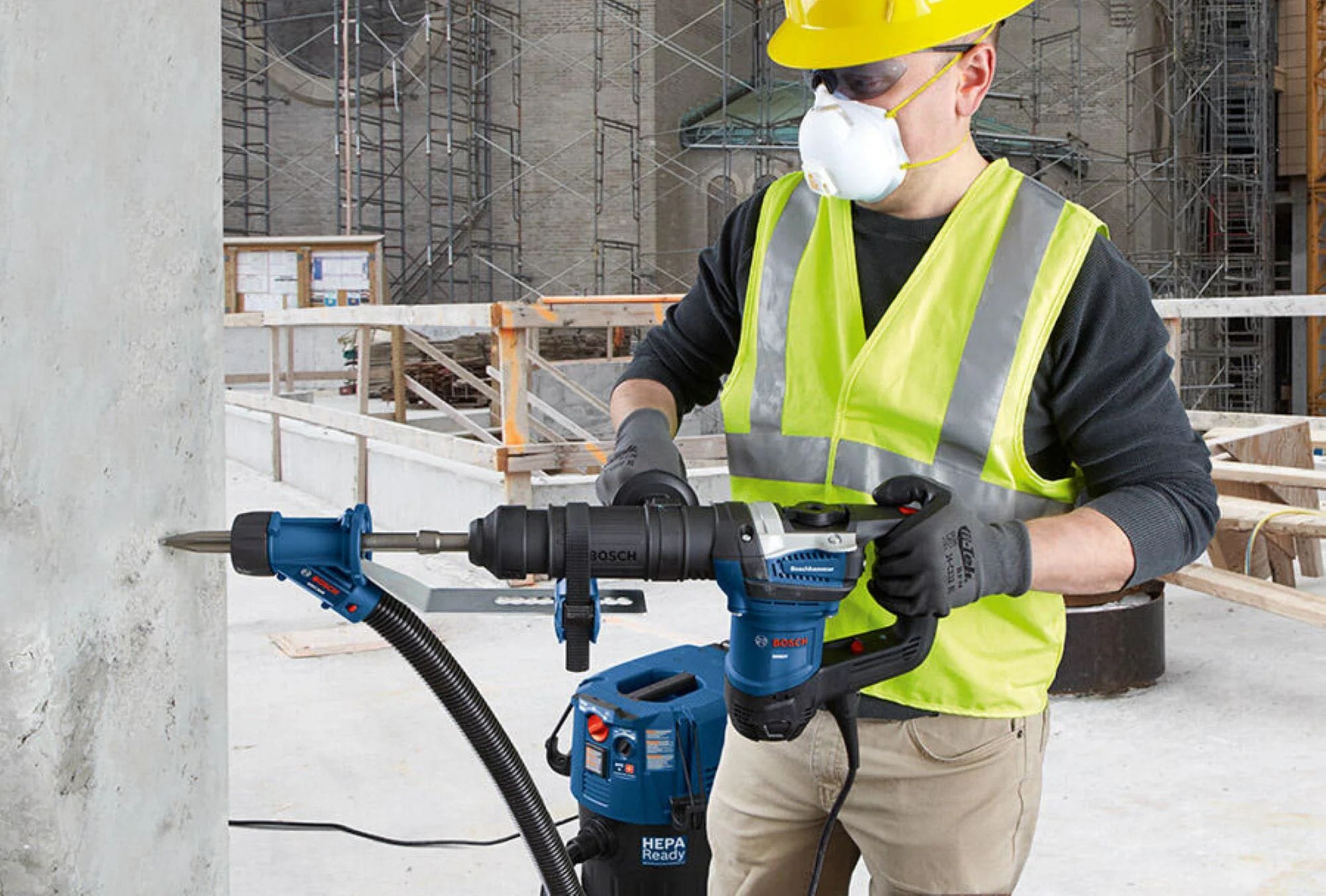 A construction professional wearing a yellow hard hat, dust safety mask, and high visibility vest using a heavy duty Bosch rotary hammer with a dust collection vacuum attachment to drive a moil point chisel bit horizontally into a concrete structure.