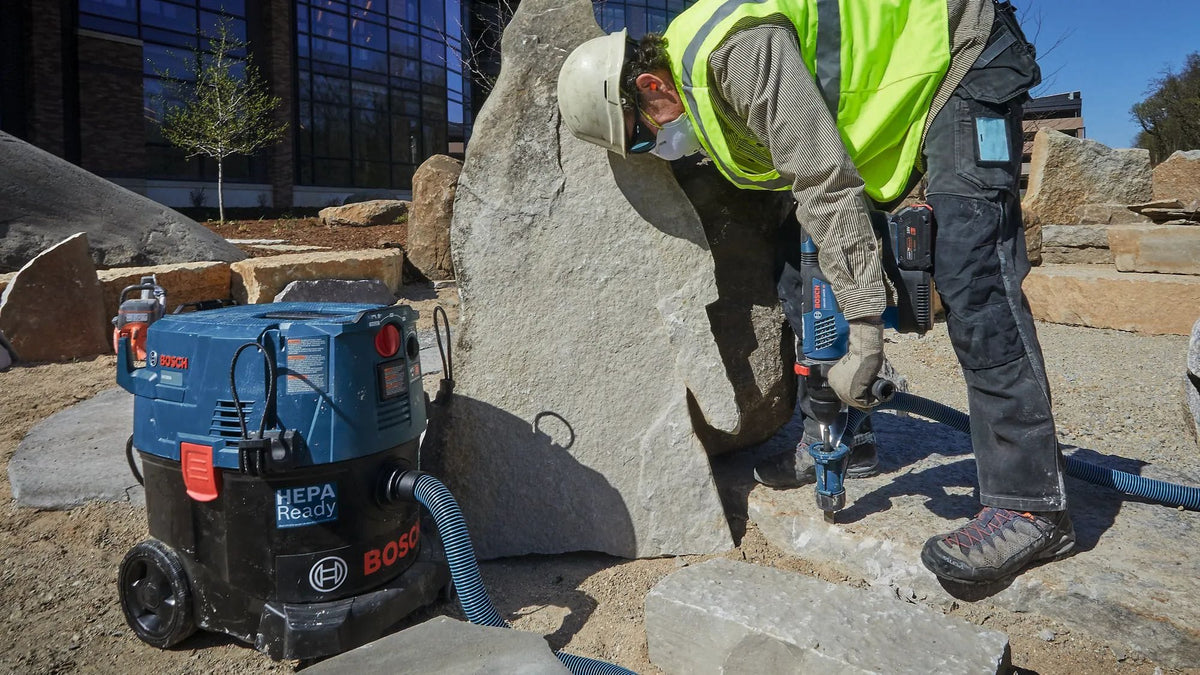 An outdoor job site worker wearing a safety helmet and high visibility gear utilizing a handheld Bosch hammer drill to chip away structural stone sections next to a Bosch industrial dust extractor vacuum unit.