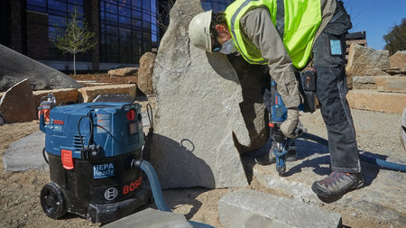 An outdoor job site worker wearing a safety helmet and high visibility gear utilizing a handheld Bosch hammer drill to chip away structural stone sections next to a Bosch industrial dust extractor vacuum unit.