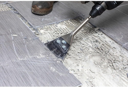 An action shot of the Bosch Bulldog floor scraper in use, demonstrating the sharp steel blade's ability to efficiently peel up ceramic floor tiles and hardened thinset from a concrete subfloor.