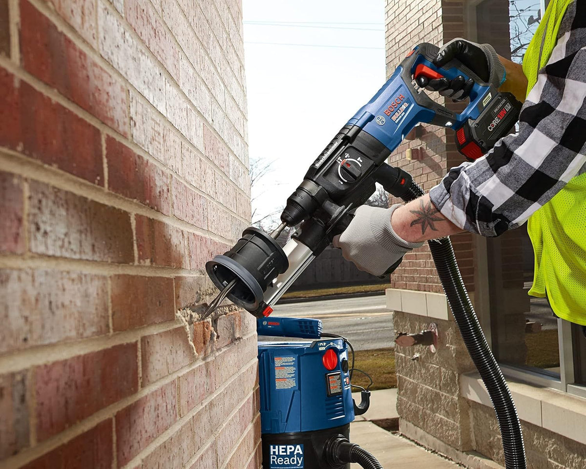 An industrial tradesman utilizing a blue cordless Bosch rotary hammer drill fitted with a dust extraction shroud assembly and HEPA ready vacuum attachment to drive a star point twist demolition chisel bit directly into a red brick masonry wall.