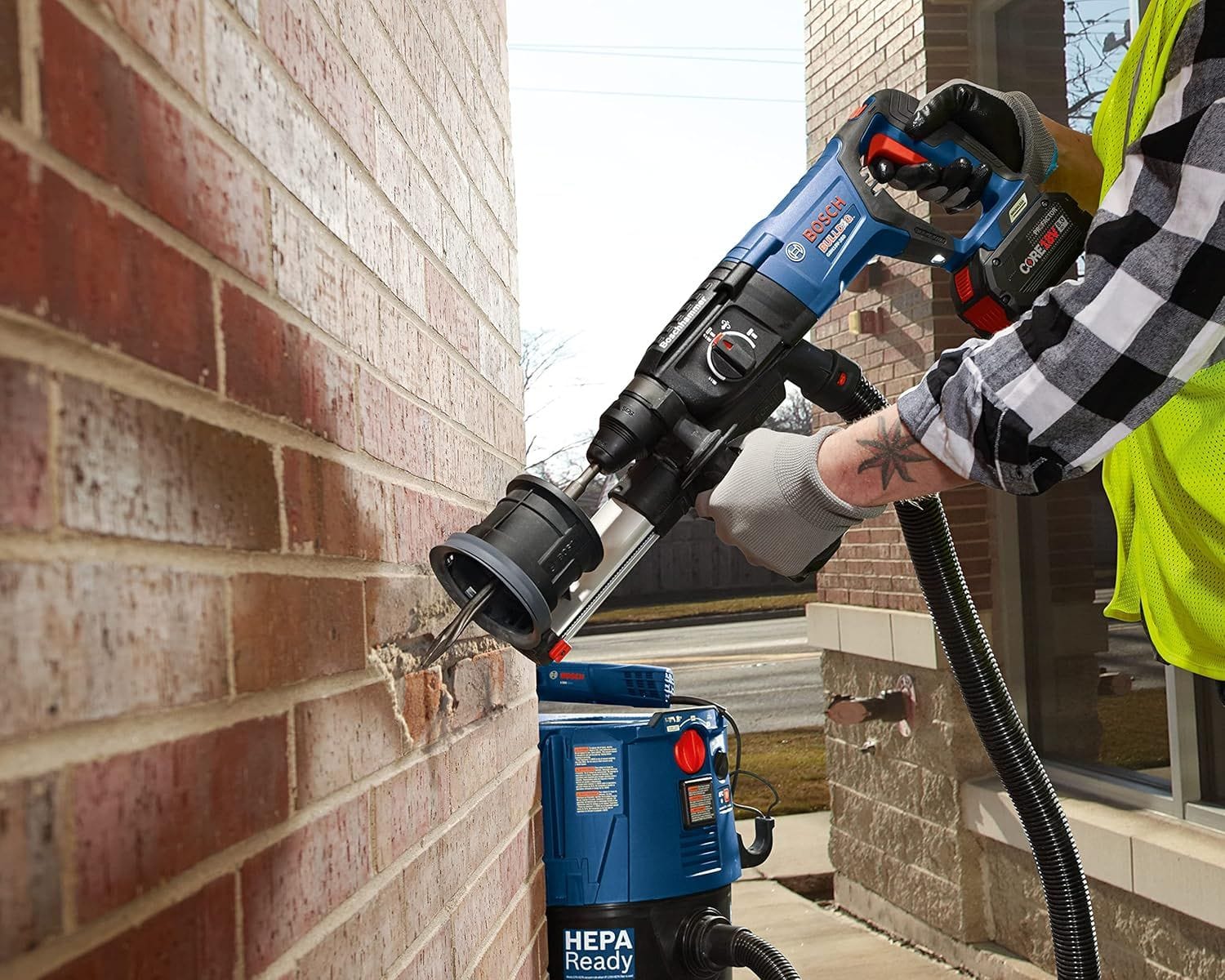 An industrial tradesman utilizing a blue cordless Bosch rotary hammer drill fitted with a dust extraction shroud assembly and HEPA ready vacuum attachment to drive a star point twist demolition chisel bit directly into a red brick masonry wall.