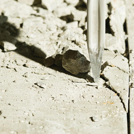 Close up high angle field shot of the star point tip profile on a Bosch self sharpening twist chisel bit initiating a stress line fracture crack inside a weathered concrete slab surface background.