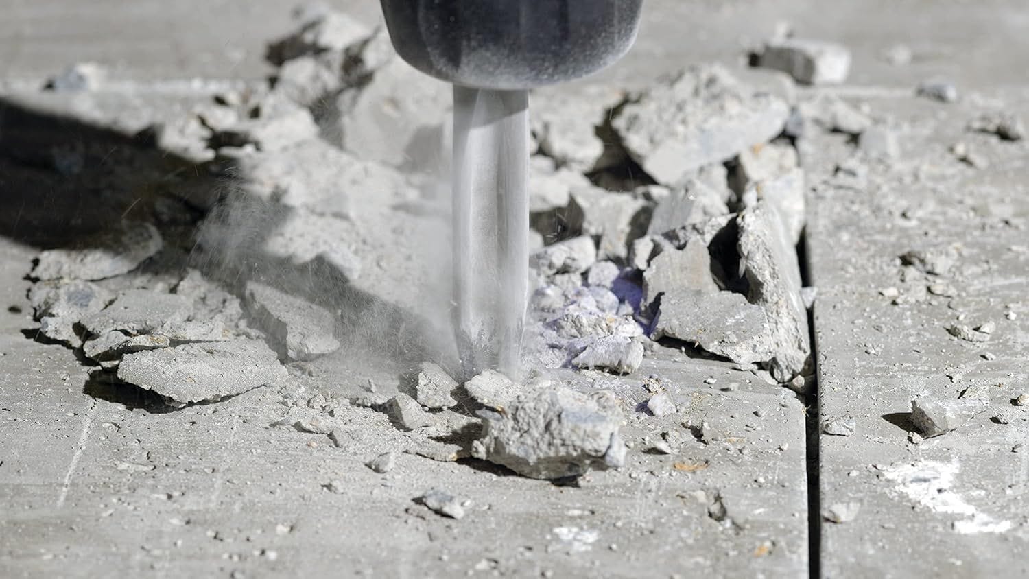 Action detail view of a heavy duty SDS plus rotary chipping hammer driving a Bosch star point twist chisel bit vertically into a cured concrete pad causing stone chips and fine masonry aggregate dust particles to erupt under impact force.