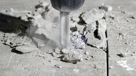 Action detail view of a heavy duty SDS plus rotary chipping hammer driving a Bosch star point twist chisel bit vertically into a cured concrete pad causing stone chips and fine masonry aggregate dust particles to erupt under impact force.