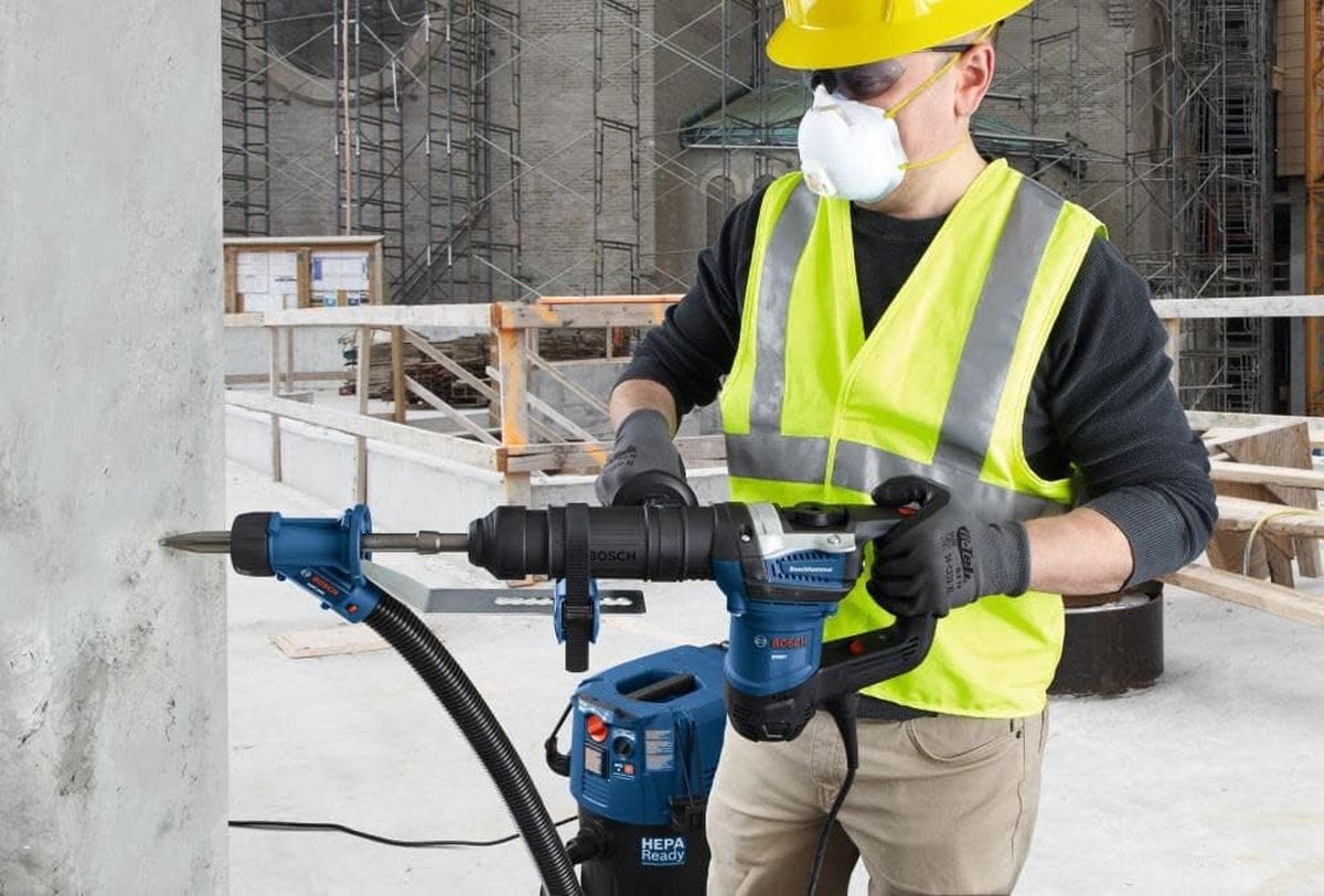 An on-site construction application photo showing an operator wearing a hard hat, safety glasses, and a dust mask using a heavy blue Bosch combination hammer drill to drive a bit horizontally into a commercial concrete structural wall, with a HEPA dust extractor vacuum vacuuming the work zone.