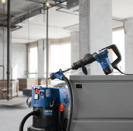 A commercial product placement photo showcasing a blue Bosch demolition hammer tool resting on top of a gray metal container box inside a concrete building under construction, paired with an active black Bosch HEPA-ready dust collection vacuum system on the floor.