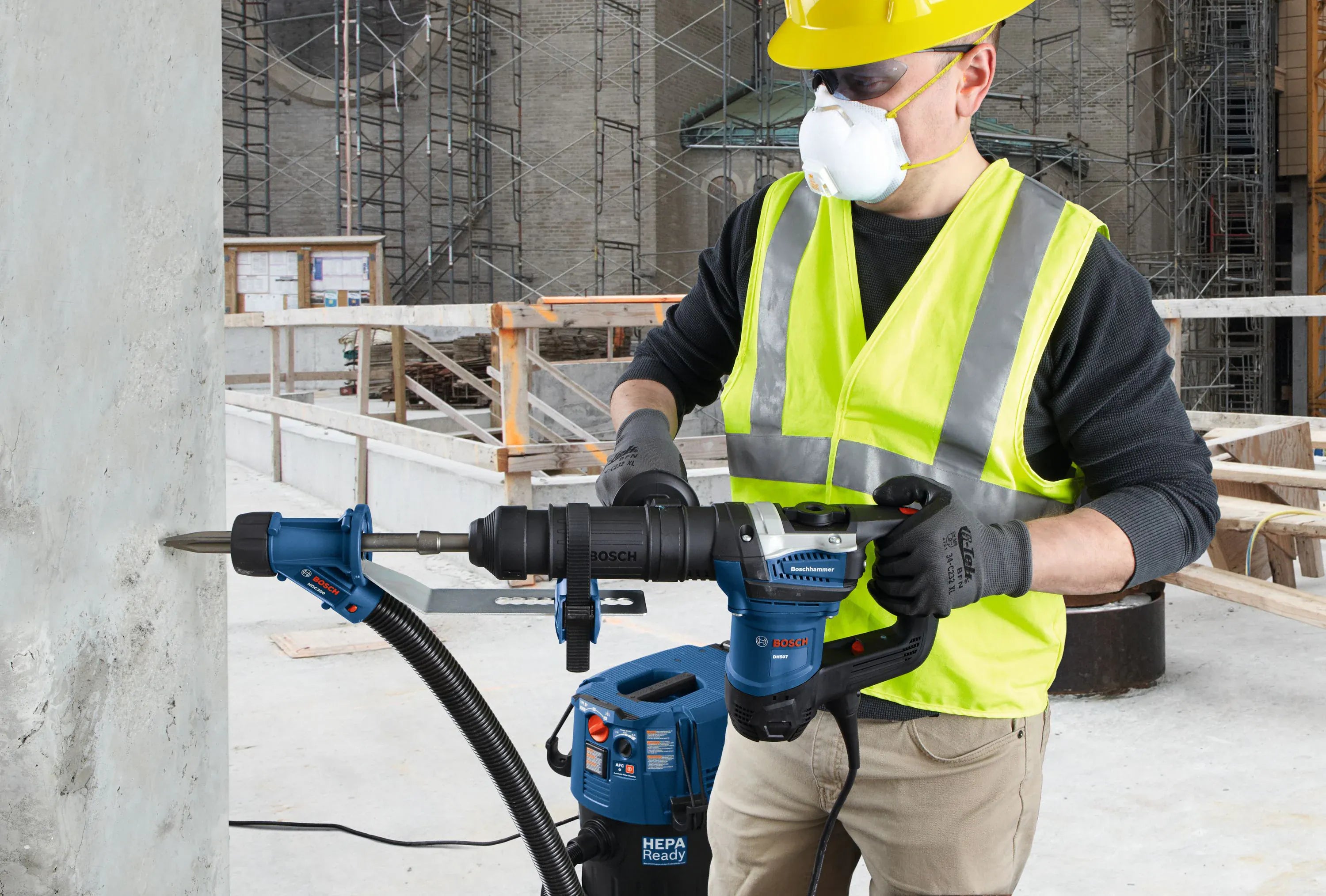 A utility contractor wearing a yellow hard hat, safety glasses, high-visibility vest, and a protective respirator mask uses an SDS-max rotary hammer drill equipped with a bull point chisel bit to punch a clean fault line into a vertical concrete structural wall, supported by an adjacent blue HEPA-ready dust extractor vacuum system.