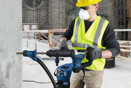 A utility contractor wearing a yellow hard hat, safety glasses, high-visibility vest, and a protective respirator mask uses an SDS-max rotary hammer drill equipped with a bull point chisel bit to punch a clean fault line into a vertical concrete structural wall, supported by an adjacent blue HEPA-ready dust extractor vacuum system.