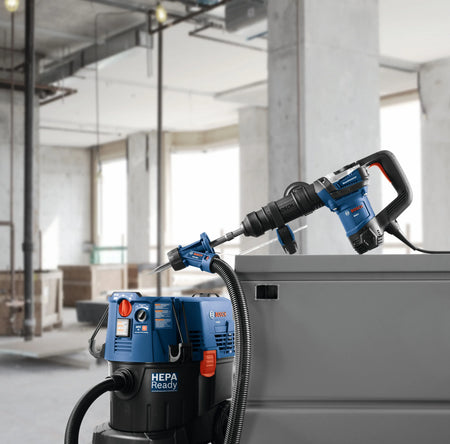 A heavy-duty blue Bosch SDS-max demolition hammer drill equipped with a dust collection shroud and a bull point bit rests balanced on a gray industrial structure layout, positioned directly above an inline blue HEPA-ready dust extractor vacuum unit within an active commercial jobsite interior.