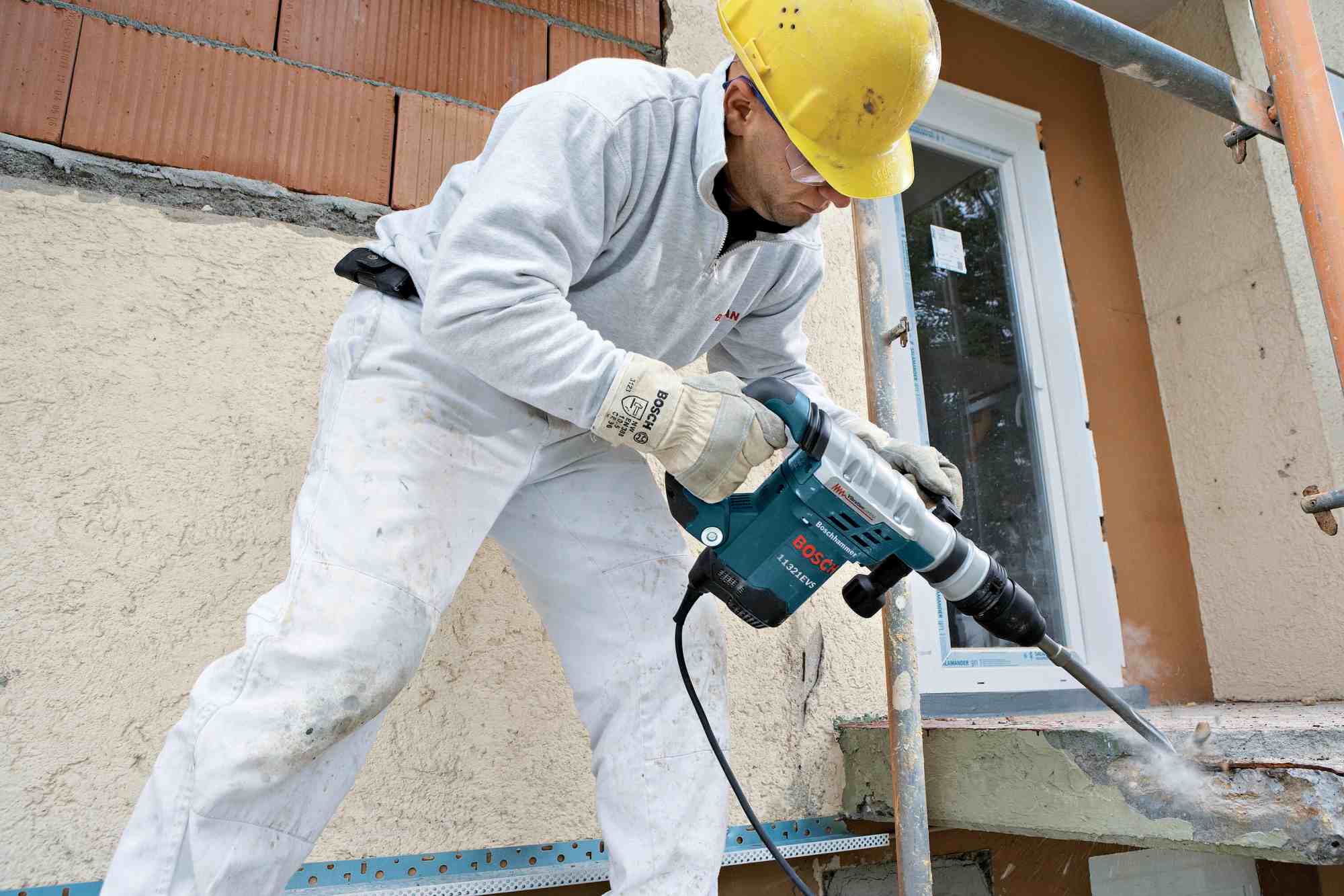 A construction professional wearing a hard hat and safety glasses stands on scaffolding, using a heavy-duty corded Bosch SDS-Max demolition hammer paired with the 16-inch R-Tec chisel to aggressively break apart a thick concrete window ledge.
