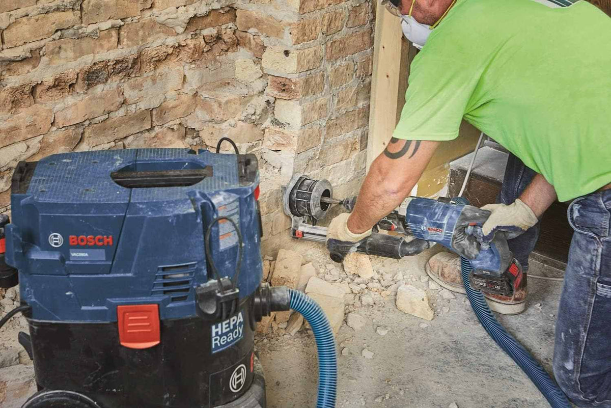 A construction worker uses the Bosch SDS-Max R-Tec demolition chisel attached to a rotary hammer with a dust shroud to break away thick brick and mortar from an exterior wall, while a Bosch HEPA ready vacuum actively collects the hazardous dust.