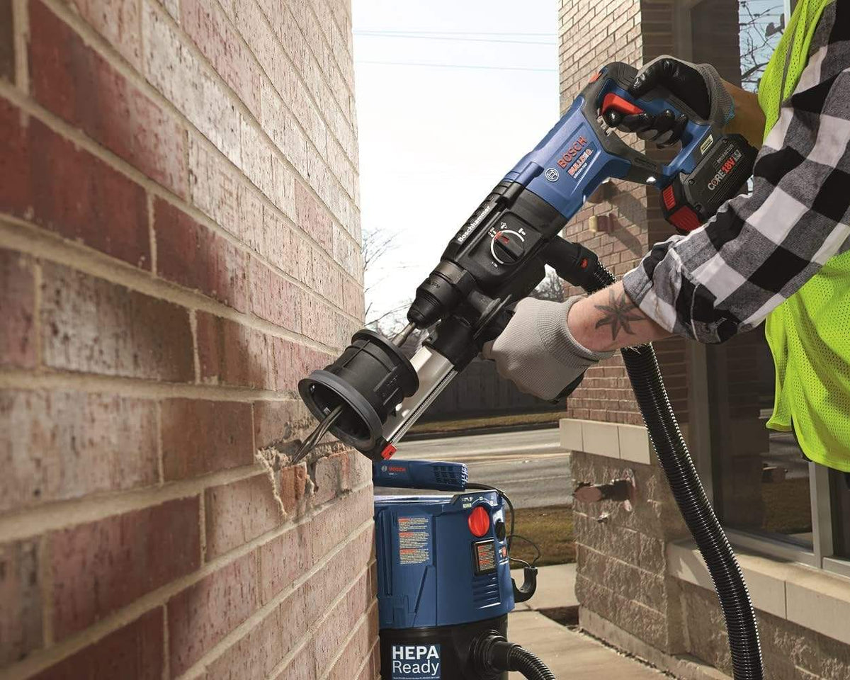 A professional safely operating a cordless Bosch SDS-Max rotary hammer equipped with an attached dust collection system uses the heavy-duty R-Tec bull point chisel to accurately penetrate and demolish a section of an outdoor red brick wall.