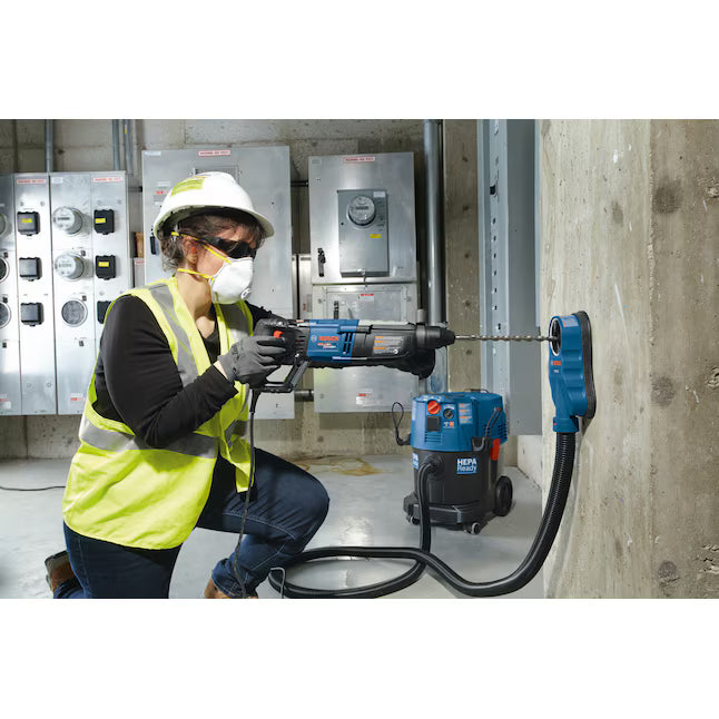 A trade professional wearing a white hard hat, high-visibility vest, and a protective respirator mask uses a corded Bosch rotary hammer drill to drill into a vertical concrete support pillar inside a utility basement, while a blue HEPA-ready dust extractor vacuum collects dust at the hole source.