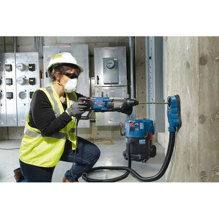A trade professional wearing a white hard hat, high-visibility vest, and a protective respirator mask uses a corded Bosch rotary hammer drill to drill into a vertical concrete support pillar inside a utility basement, while a blue HEPA-ready dust extractor vacuum collects dust at the hole source.