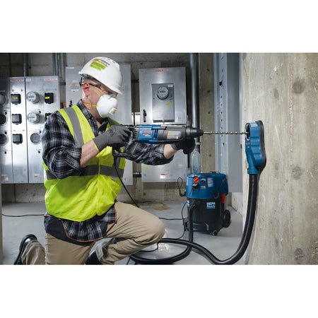 A construction worker kneeling in an electrical room operating a corded professional Bosch SDS rotary hammer drill to bore a clean anchor point into a reinforced concrete wall, utilizing an adjacent blue dust extractor vacuum for on-site OSHA Table 1 compliance.