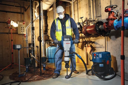 A utility contractor wearing a hard hat, safety glasses, high-visibility safety vest, and a protective respirator mask operates a heavy-duty Bosch Brute Turbo electric breaker hammer inside a commercial boiler room, utilizing a vacuum hose routing system connected to a blue HEPA-ready dust extractor vacuum for on-site OSHA Table 1 compliance.
