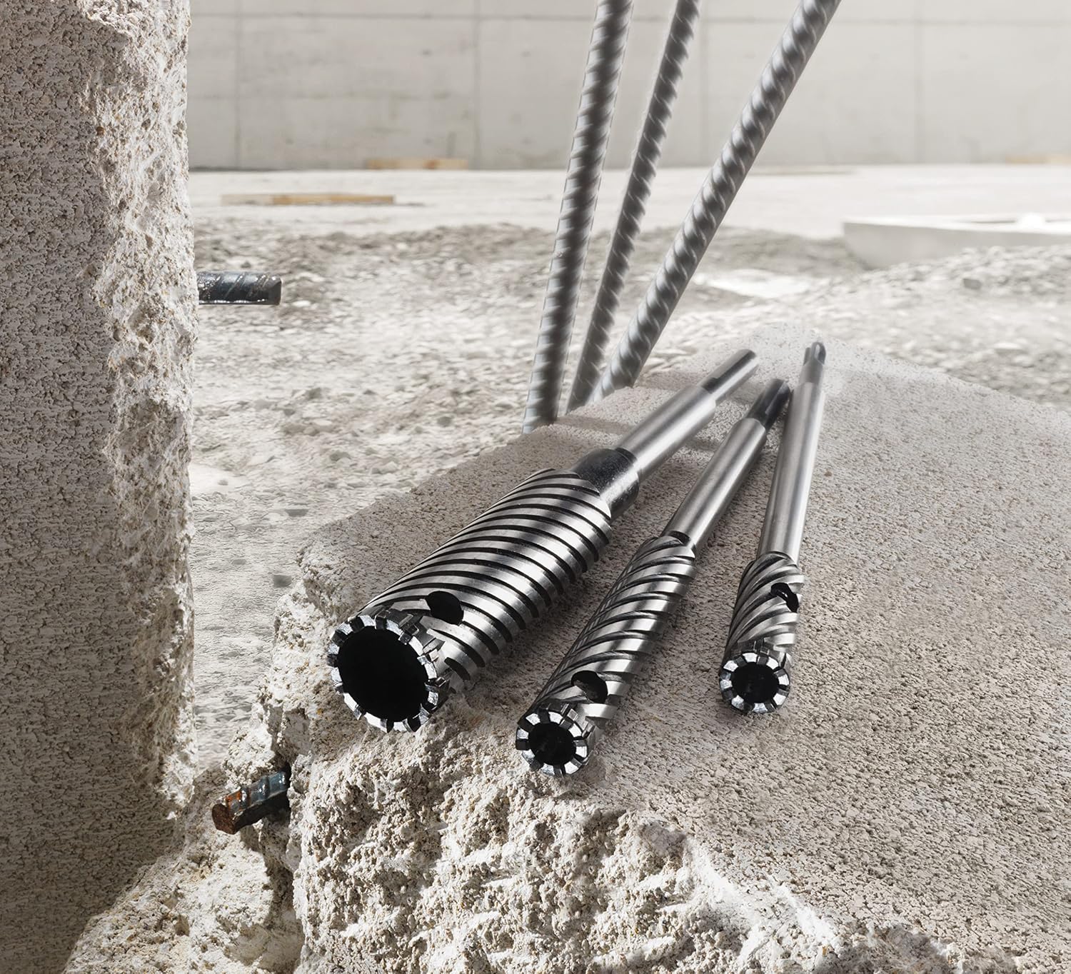 An on-site construction application shot showcasing three different sizes of heavy-duty Bosch rebar cutter bits resting flat on a textured concrete slab, detailing their wide cylindrical bodies, deep-cut steep spiral flutes, ventilation relief holes, and teeth-lined carbide cutting rings against a background of exposed steel rebar rods.