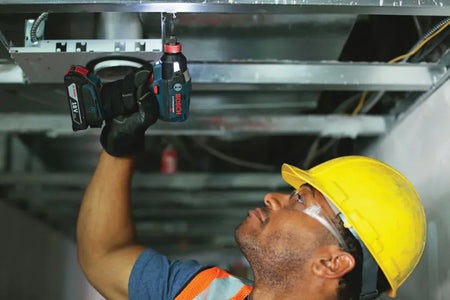 A professional electrician wearing a hard hat and safety glasses uses the Bosch 18V two-in-one impact driver to install electrical fixtures into overhead metal framing, showcasing the tool's compact ergonomics and reach.