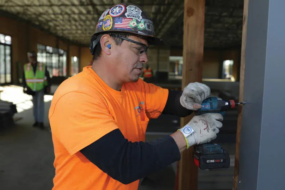 A professional metal worker using the Bosch 18V two-in-one impact driver to fasten a self-tapping screw into a structural steel beam on a commercial construction site.