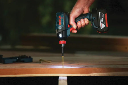 A jobsite application shot showing the Bosch 18V brushless impact driver driving a long wood screw into structural lumber, highlighting the integrated three-LED light ring for shadow-free illumination of the workspace.