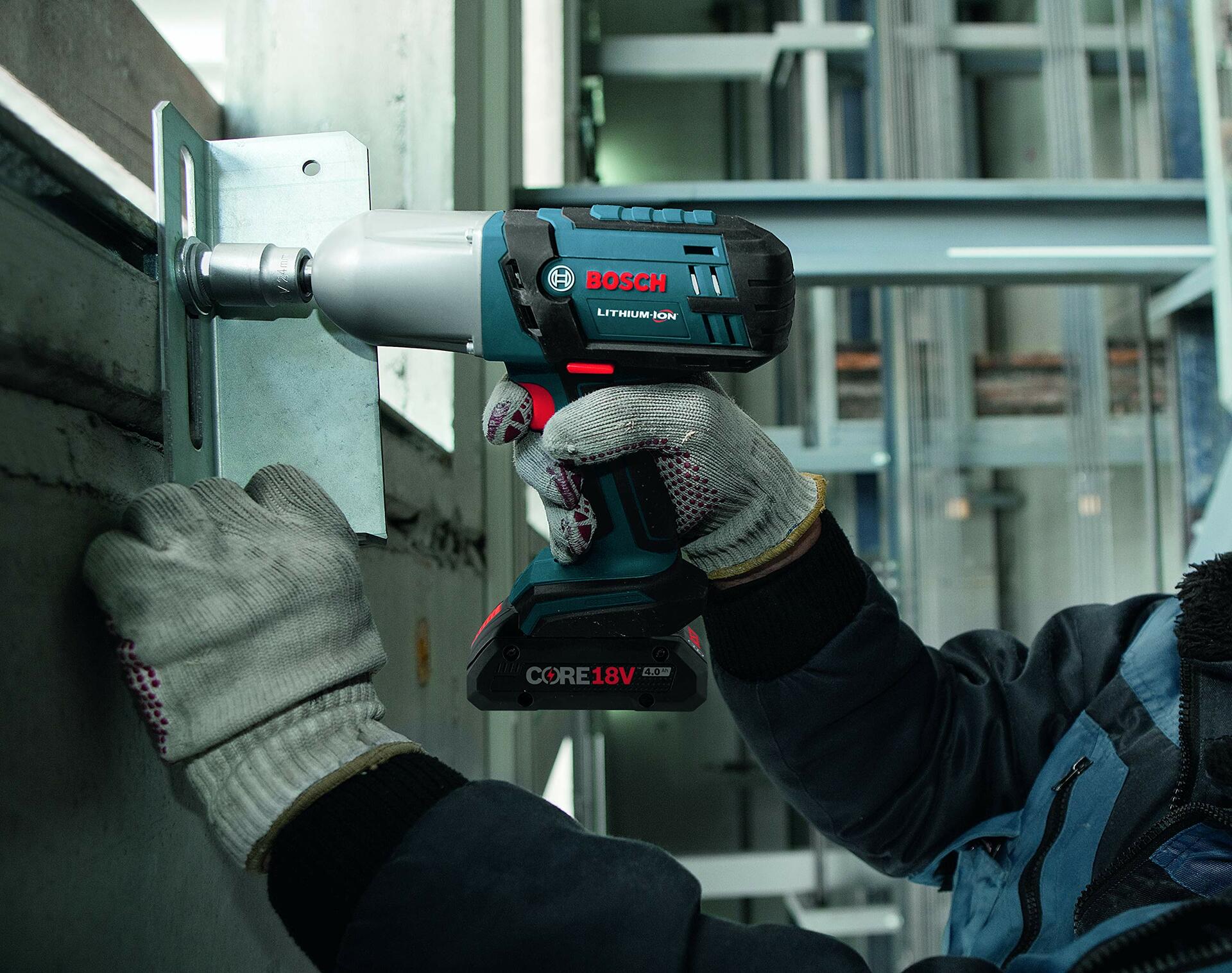A worker in gloves uses the Bosch IWHT180 18V impact wrench with its LED light on to tighten a large bolt on a metal bracket in an industrial setting.