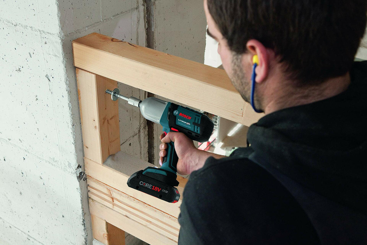 A worker with ear protection uses the Bosch IWHT180 18V impact wrench with a long extension to drive a large fastener into a wooden frame against a concrete wall.