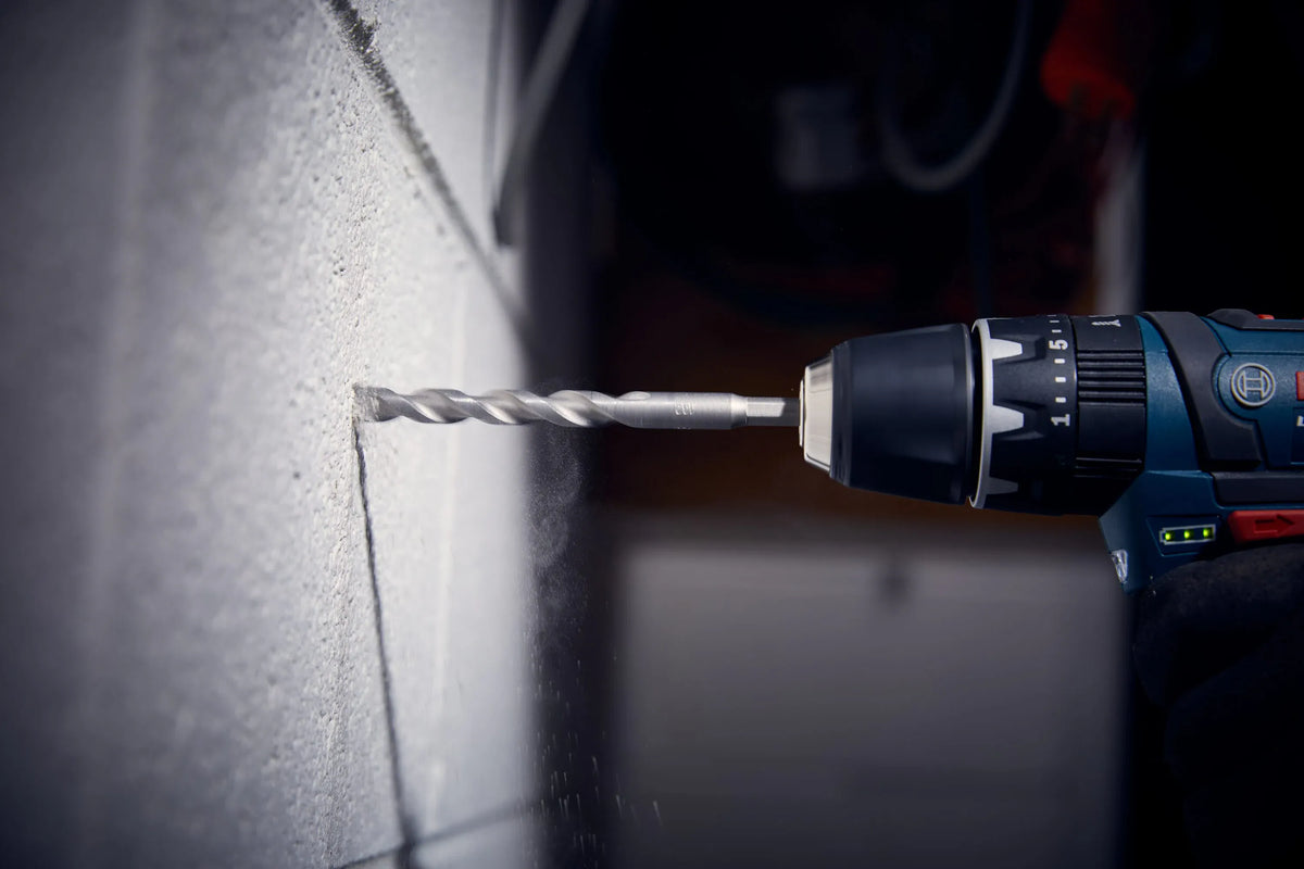 A close-up action view of a professional technician using a cordless hammer drill tool to bore a clean fastener hole horizontally into an interior painted white concrete block wall surface. The tool is equipped with a premium carbide-tipped percussion bit from the Bosch LBHXS7U hex shank set, capturing fine masonry dust actively expelling from the deep-cut spiral flutes.