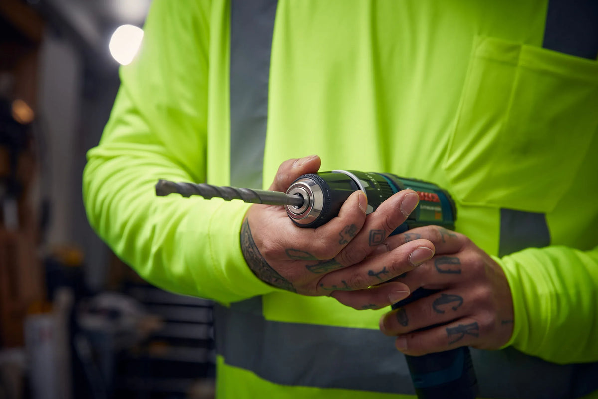 A close-up real-world workspace view of a construction tradesman wearing a high-visibility yellow safety shirt checking tool setups. The worker's tattooed hands are shown loosening or tightening the metal keyless chuck collar on a cordless drill driver to secure a silver hex shank masonry bit from the LBHXS7U percussion kit.