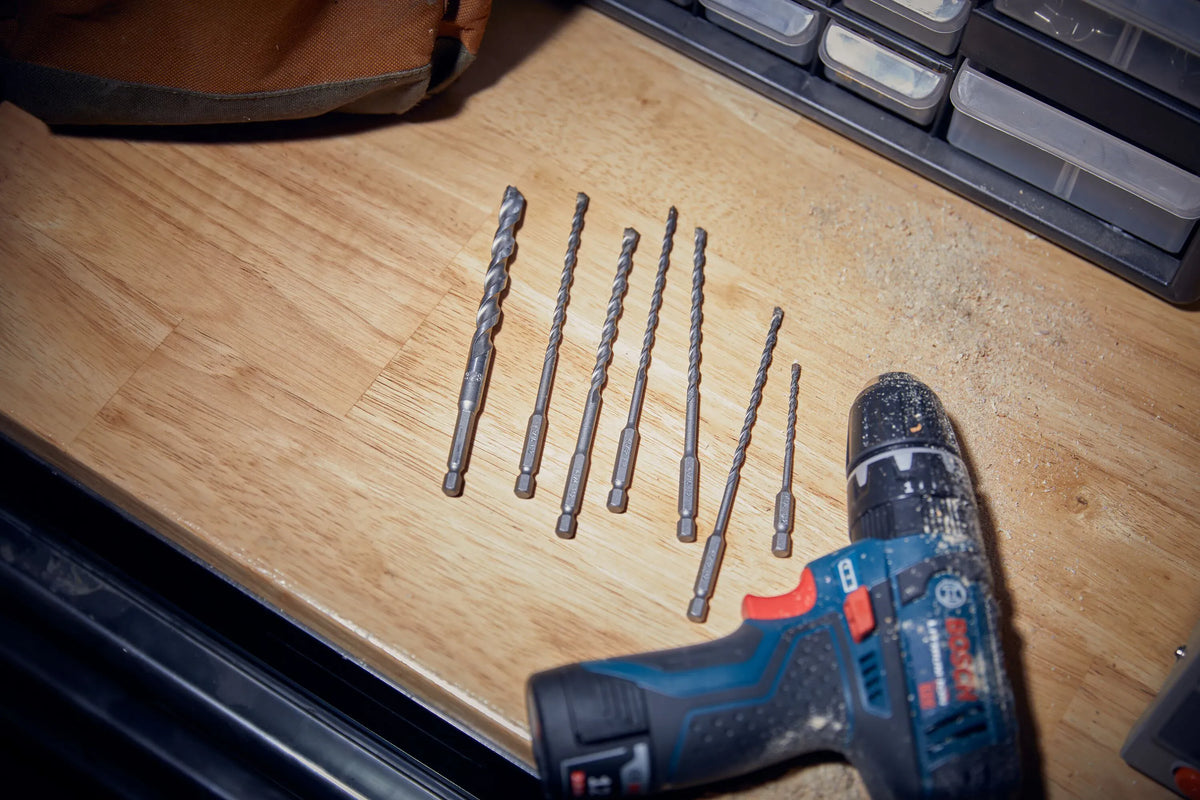 A workspace layout image capturing the complete loose contents of the Bosch LBHXS7U 7-piece masonry bit set fanned out horizontally on a smooth wooden workshop benchtop. Sited next to a loose brown canvas tool pouch and an unattached blue Bosch cordless hammer drill driver, the image shows the diverse selection of impact-rated 1/4-inch hex driven masonry accessories ready for field operation.