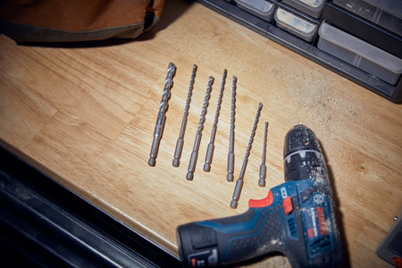 A workspace layout image capturing the complete loose contents of the Bosch LBHXS7U 7-piece masonry bit set fanned out horizontally on a smooth wooden workshop benchtop. Sited next to a loose brown canvas tool pouch and an unattached blue Bosch cordless hammer drill driver, the image shows the diverse selection of impact-rated 1/4-inch hex driven masonry accessories ready for field operation.
