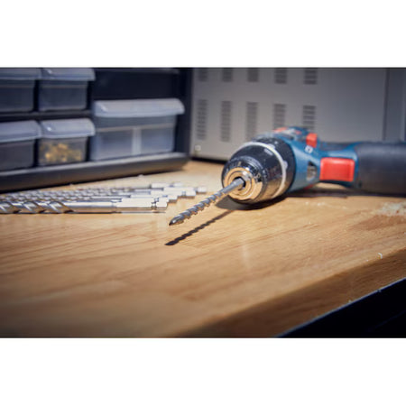 A low-angle workshop perspective shot focusing on a silver hex shank masonry drill bit locked into the keyless chuck of a blue Bosch cordless power drill driver resting on a wooden workbench. The shallow depth-of-field blur highlights the specialized diamond-ground carbide tip configuration in the foreground while the remaining loose bits from the LBHXS7U kit wait on the table deck behind it.