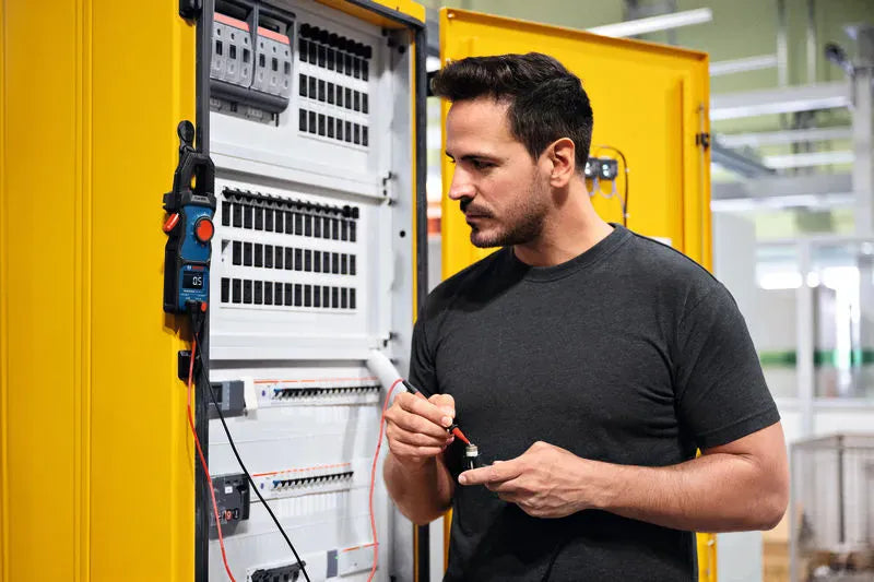 A professional electrician working at a bright yellow industrial machinery panel box utilizing the Bosch MH1 multi-meter mounting system. The industrial-grade magnet firmly grips the metallic cabinet corner frame, suspending a digital clamp meter hands-free to optimize user safety and manual testing dexterity during complex terminal maintenance.