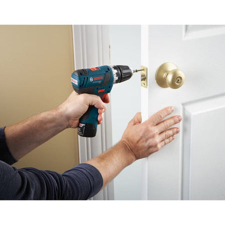 A person using a cordless drill and a driver bit from the Bosch MS4034 set to install a screw into a brass door latch plate on a white interior door.
