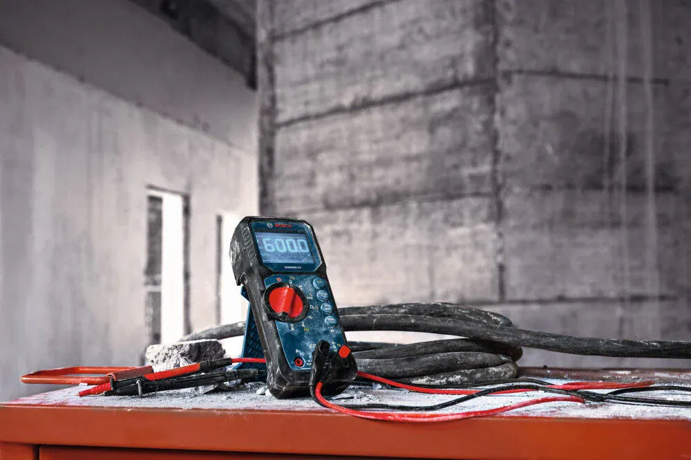 The Bosch MS90 double-insulated silicone test lead wire set shown in a real-world application, plugged into a rugged digital multimeter positioned on a dusty, concrete jobsite workbench. The flexible red and black electrical testing cables are laid out across the work surface near heavy industrial wiring, showing off their durability under demanding field diagnostic and troubleshooting conditions.