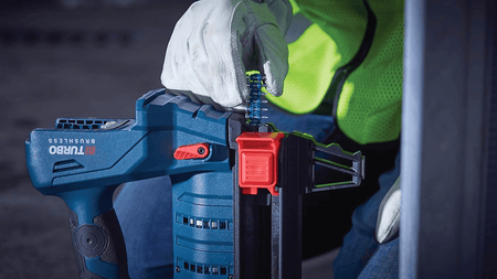 A real-world commercial framing application shot showing a construction worker wearing a white leather glove inserting a blue collated strip of concrete nails into the magazine feeder mechanism of a cordless blue Bosch Biturbo brushless concrete nailer tool.