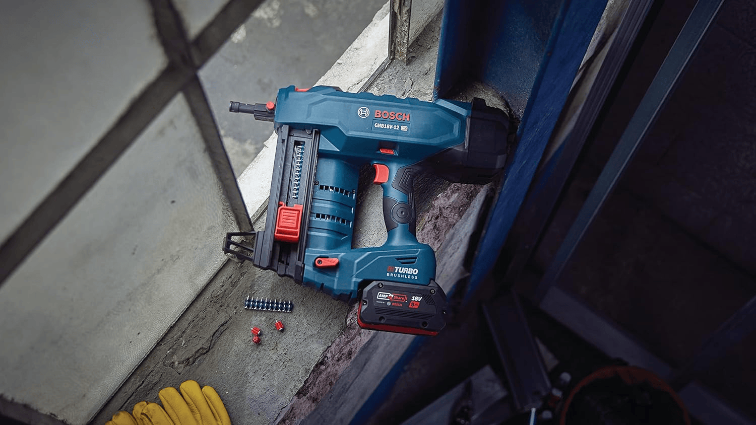 A jobsite lifestyle photograph showing a blue Bosch GNB18V-12 18V concrete nailer resting on a concrete ledger next to a detached strip of collated track pins and a pair of yellow leather work gloves near an industrial window frame.
