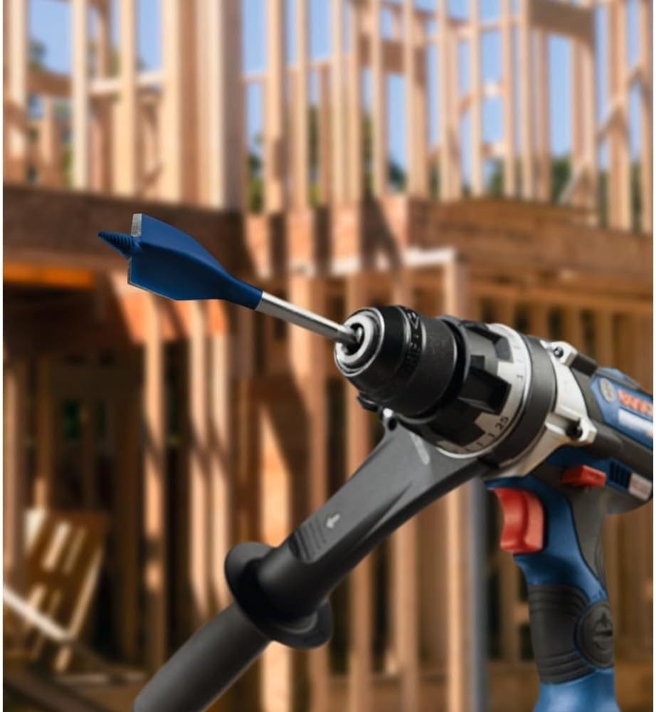 A close-up of a Bosch Nail Strike spade bit mounted in a professional drill driver, set against a blurred background of a residential wood-frame house under construction.