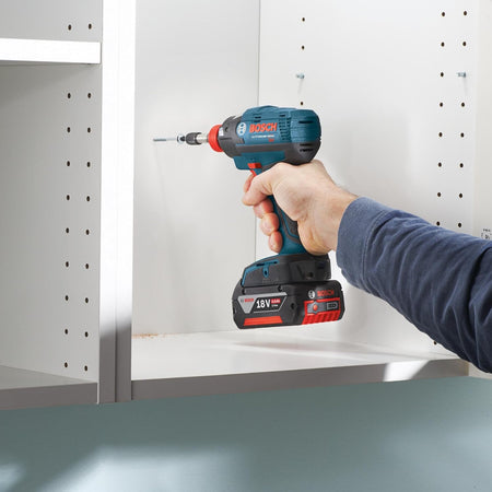 An interior application shot showing a technician assembling white melamine cabinetry units, utilizing a handheld blue Bosch impact drill with a long Phillips screwdriver bit to anchor a fastening screw into pre-drilled side panels.