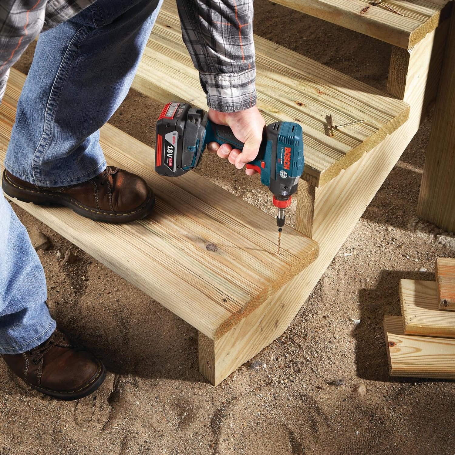 A jobsite photograph of a contractor installing pressure-treated wood stairs, using a cordless Bosch 18V impact driver with an extended 3-1/2 inch Phillips power bit to drive heavy-duty deck screws straight down into the step treads.