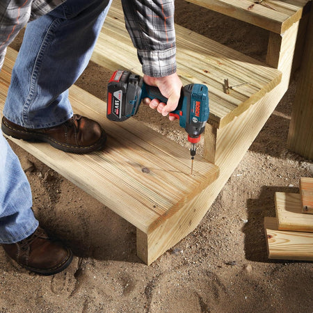 A jobsite photograph of a contractor installing pressure-treated wood stairs, using a cordless Bosch 18V impact driver with an extended 3-1/2 inch Phillips power bit to drive heavy-duty deck screws straight down into the step treads.
