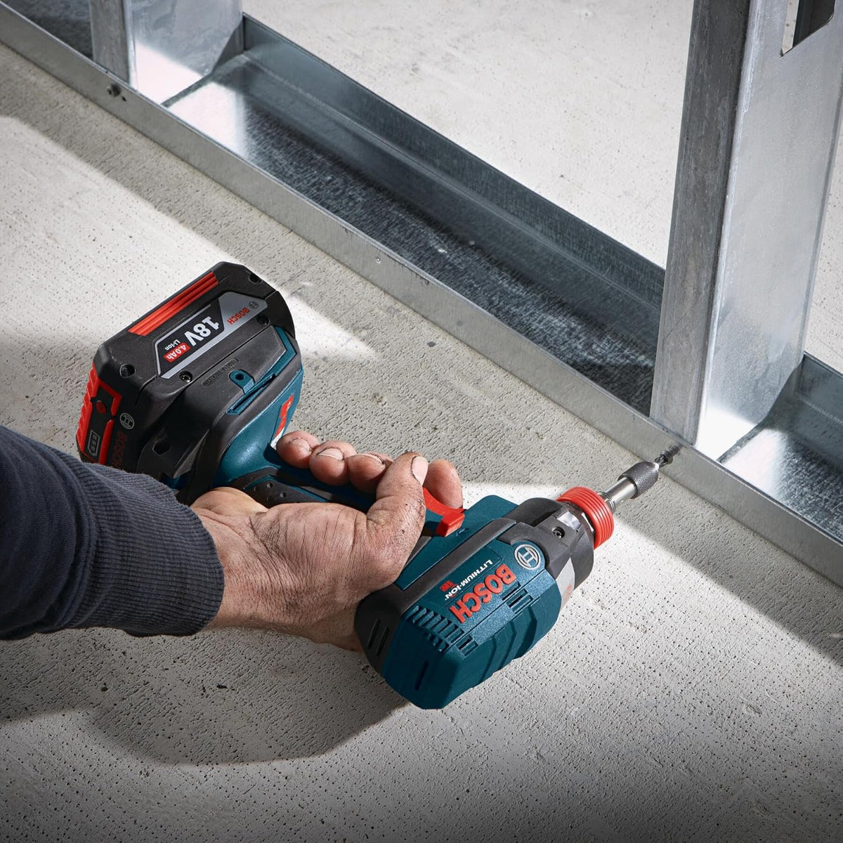 An on-site construction application shot of an operator using a blue Bosch 18V cordless impact driver equipped with a 3-1/2 inch Phillips #2 power bit to drive a structural screw directly through a galvanized steel wall stud.