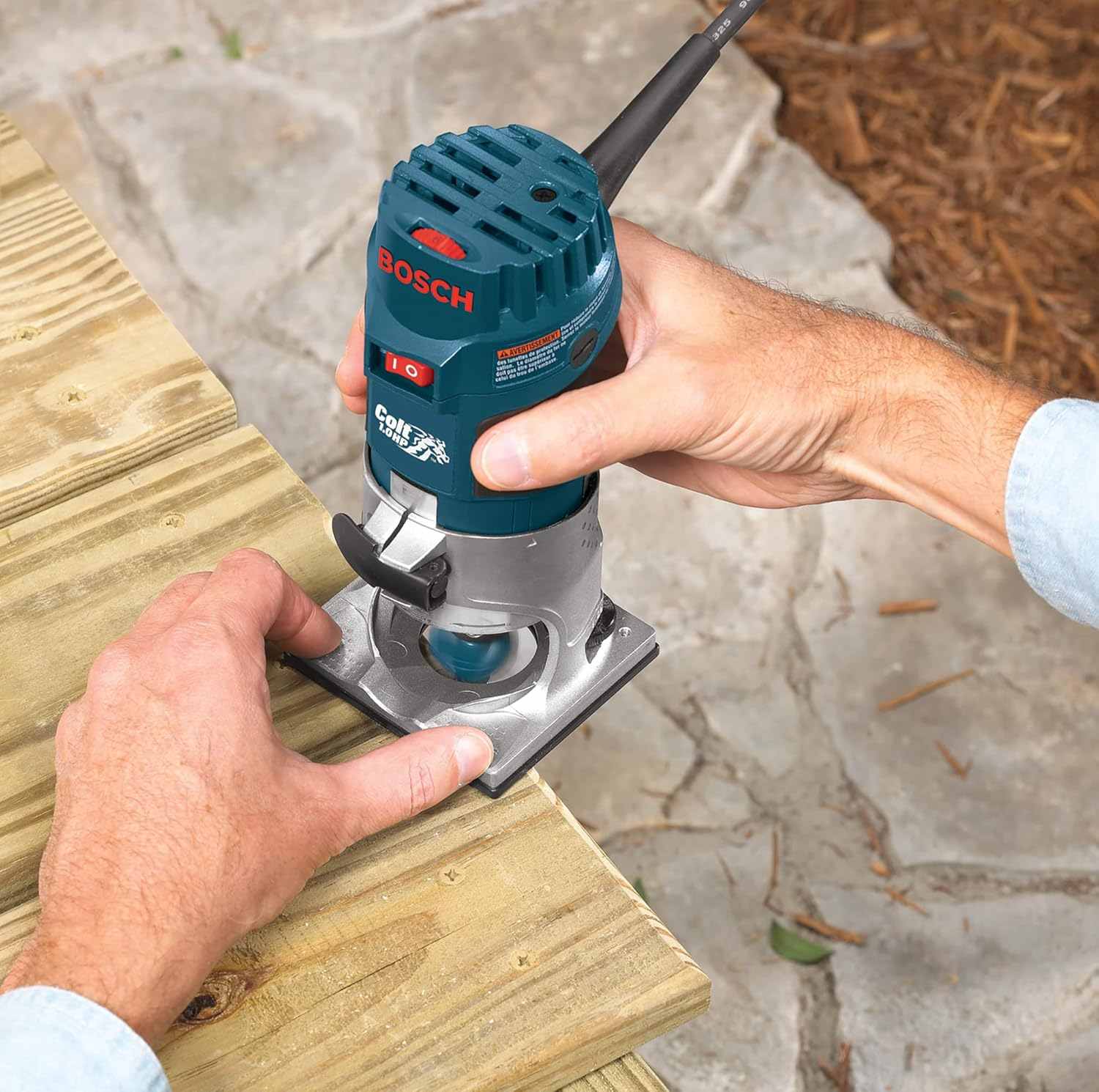 A high-angle shot of a user guiding the palm router along the edge of a wooden deck board to create a rounded edge profile.