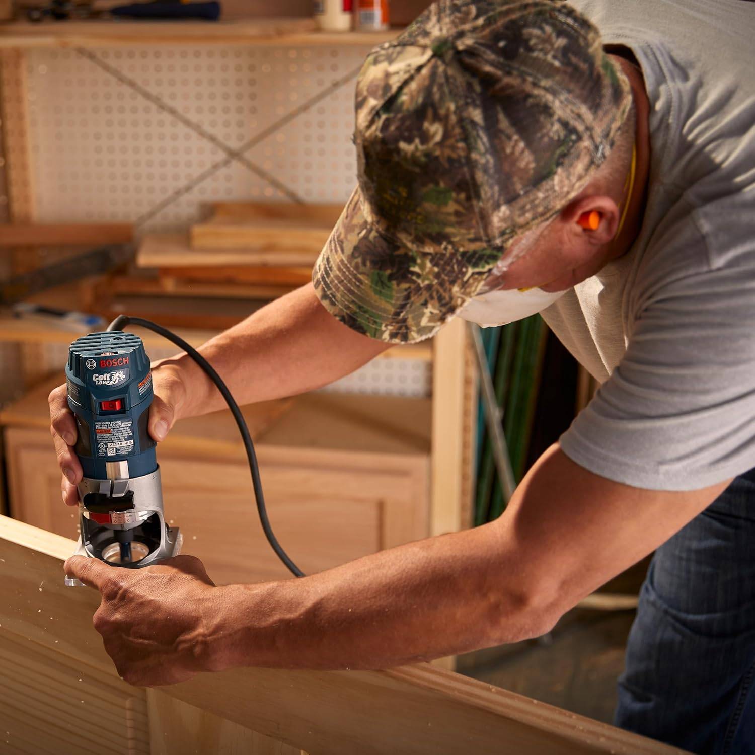 A woodworker wearing safety glasses using the router to shape the edge of a wooden frame in a workshop setting.