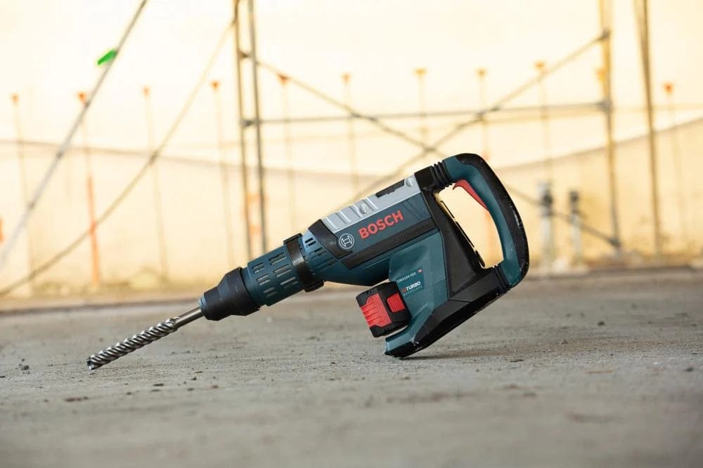 The Bosch PROFACTOR 18V Hitman SDS-max brushless cordless rotary hammer resting on a concrete floor surface with a long masonry drill bit attached, ready for heavy-duty commercial drilling tasks.