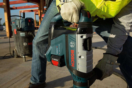 A construction worker wearing protective leather gloves and a high-visibility vest carrying the portable Bosch PROFACTOR 18V Hitman cordless rotary hammer across a commercial concrete job site.