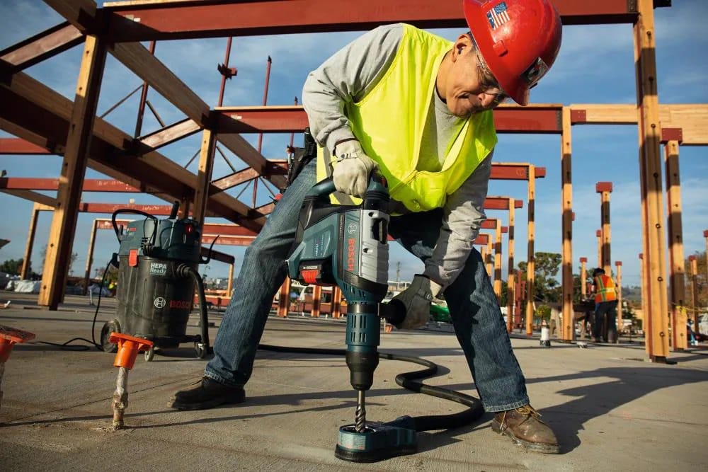 A contractor using the Bosch PROFACTOR 18V Hitman SDS-max rotary hammer to drill vertically into a concrete slab, paired with a dust extraction vacuum attachment for a clean workspace.