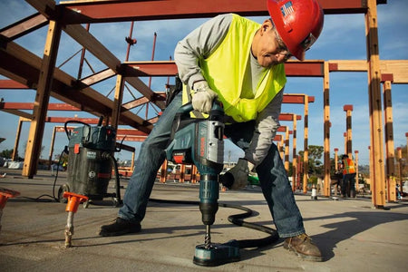 A contractor using the Bosch PROFACTOR 18V Hitman SDS-max rotary hammer to drill vertically into a concrete slab, paired with a dust extraction vacuum attachment for a clean workspace.