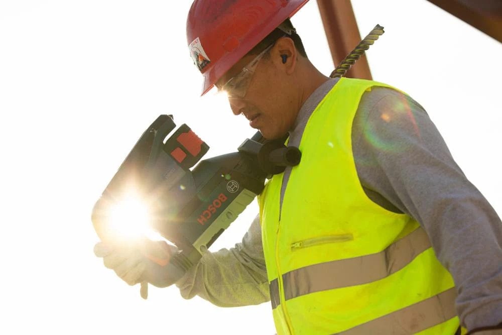 A job site worker safely carrying the highly portable Bosch PROFACTOR 18V Hitman cordless rotary hammer over his shoulder while wearing a hard hat and safety glasses on a sunny construction site.