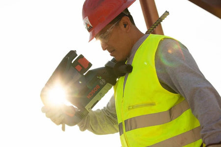 A job site worker safely carrying the highly portable Bosch PROFACTOR 18V Hitman cordless rotary hammer over his shoulder while wearing a hard hat and safety glasses on a sunny construction site.